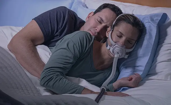 Un couple dormant blotti l'un contre l'autre dans son lit, la femme portant un masque AirFit F40 de ResMed.