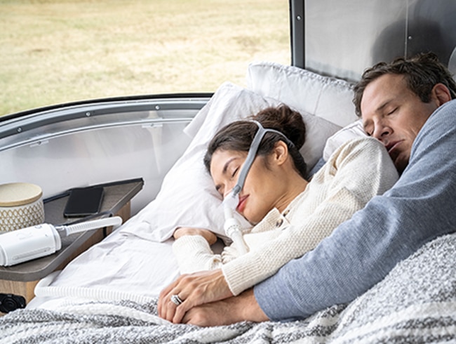  Personne en train de dormir dans un lit avec un appareil PPC compact posé à proximité, illustrant l’utilisation de la thérapie pendant un voyage en van.