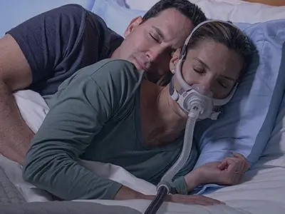 Un couple dormant blotti l'un contre l'autre dans son lit, la femme portant un masque AirFit F40 de ResMed.